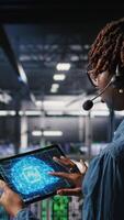 Vertical Data center worker on industrial platform speaks in headset microphone while overseeing maintenance on AI rigs below. Admin uses tablet to coordinate deep learning system tasks with team, camera A video