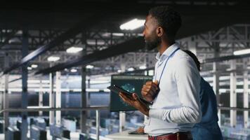 Technician walks in data center, doing maintenance tasks on tablet, ensuring optimal performance. IT expert reviewing diagnostics and applying patches to server hub gear, improving stability, camera B video