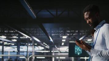 Engineer walking on data center industrial platform, performing system maintenance. IT professional using tablet to do upgrades, resolving bugs and enhancing infrastructure stability, camera A video