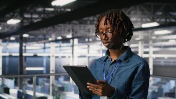 Server room admin using tablet and headset microphone to provide infrastructure tech support. Data center IT specialist resolves backend issues while coordinating with remote teams, camera B video