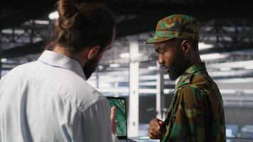 Military experts in data center reviewing drone surveillance feed on laptop screen. Army units in high tech server room doing battlefield reconnaissance operations using notebook, camera B video