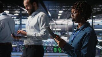 Data center engineer wearing headset microphone providing remote diagnostics using tablet. Technician monitors system health and guides users through infrastructure troubleshooting steps, camera A video