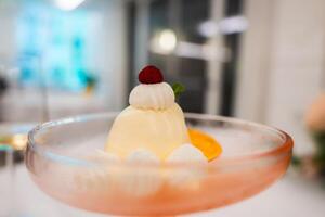 A creamy dessert in a glass dish topped with whipped cream, a red berry, and mint, surrounded by cream and orange garnish in a refined setting. photo