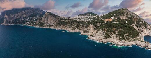 Capri Island's rugged cliffs meet the Tyrrhenian Sea, with whitewashed villas on green hillsides under a vibrant sky with soft clouds. photo