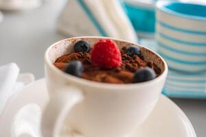 A white cup of tiramisu topped with cocoa powder, a raspberry, and blueberries on a saucer. Blurred background includes a striped cup and napkins. photo