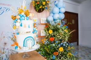 A multi tiered cake with blue, orange, and gold decorations, surrounded by floral arrangements and balloons, set in an indoor festive setting. photo