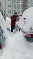 A man clearing his car of snow in defocus in the background. Snow drifts and storms. The driver in a backpack with a brush clears snow from the car standing on video