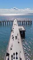 Sunny Day on a Scenic Ocean Pier. Aerial view of a pier extending into the sparkling ocean under a bright blue sky, with people strolling and relaxing. video