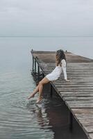 unrecognizable woman in a white dress, sitting on the edge of a wooden pier, dipping her feet into calm water with a light splash, gazing at the horizon under a soft cloudy sky, relaxation, travel photo