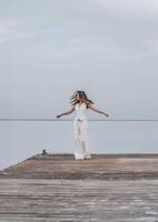 woman dancing barefoot on a wooden pier, long hair flowing as she twirls freely under a pale sky, surrounded by calm water, joyful expression in motion, freedom, wellness, motion blur photo