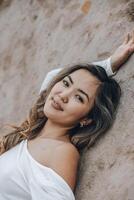 young Asian woman on beige stone wall, smiling gently in white off-shoulder top with relaxed expression, concept of beauty, natural skincare, feminine lifestyle photo