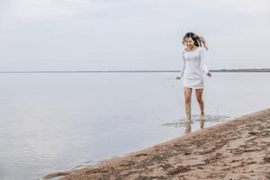 Asian woman in white dress splashing in shallow water at beach, smile, looks down, calm lake with soft horizon under cloudy sky, summer lifestyle, travel, wellness retreats, motion blur photo