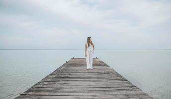 Asian woman standing barefoot on a wooden pier, gazing into the distance over a calm sea horizon, dressed in a white outfit under a pale cloudy sky, mindfulness, wellness retreat, travel blog photo