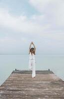 Asian woman stands barefoot with hands up on wooden pier, gazing into distance over calm sea horizon, in a white outfit under a pale cloudy sky, mindfulness, wellness retreat, travel blog photo
