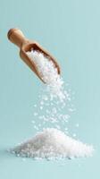 Coarse salt pours from wooden scoop onto pile of white salt crystals on light blue background creating fresh natural feel photo