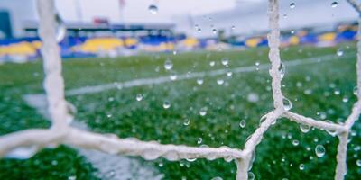 Close-up view of a soccer goal net with droplets of rain clinging to the mesh. The blurred green grass and distant stadium seating create a dynamic backdrop. photo