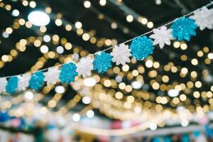 A string of glittery snowflake decorations in blue and white hangs against a backdrop of softly glowing, out-of-focus warm lights, creating a festive ambiance. photo