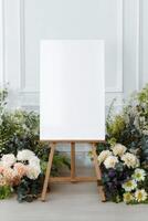 Wooden easel with a blank white canvas, surrounded by flowers in a bright studio space with a decorative white wall, awaiting its art. photo
