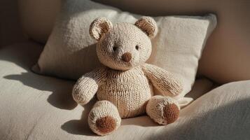 A soft, knitted teddy bear sits on a light-colored sofa next to a plush pillow. The bear has a beige body with brown accents on its ears and paws. photo