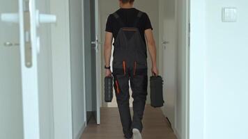 Professional repairman in overalls walks down hallway carrying toolboxes in both hands. Worker leaving bright modern interior after work. Concept of home repair or renovation video