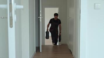 Professional repairman in overalls walks down hallway carrying toolboxes in both hands. Worker entering bright modern interior. Concept of home repair or renovation video