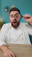 Freelancer man holds magnifying glass near face looking at camera with big zoomed eye, analyzing video