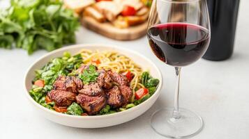 A bowl of pasta with meat and vegetables and a glass of red wine photo