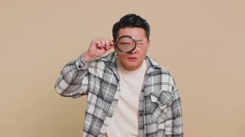 Asian mature man holds magnifying glass near face looking at camera with big zoomed eye, analyzing video