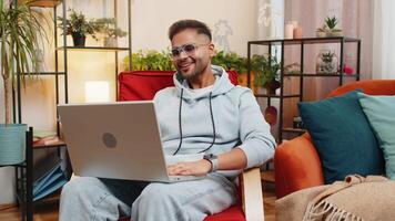 Indian man sitting on home chair, looking at laptop, making conference call with friends video