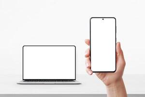 Smartphone in hand with blank screen and laptop in background on desk, both ready for UI or branding mockup photo