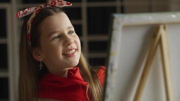 A smiling young artist in a red sweater gazes at her painting on an easel. With a creative headband, she seems content in her creative space, creating an atmosphere of focus. video