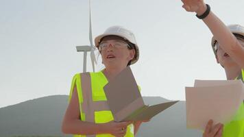 Two excited children in colorful vests examine blueprints by a tall wind turbine, showcasing teamwork and curiosity in a vibrant renewable energy setting, reflecting their passion for exploration video