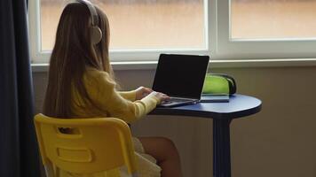 A young girl is sitting at a desk, using a laptop and wearing headphones. She is in an indoor setting by a window, engaged in learning or entertainment on her computer. The scene is bright and focused. video