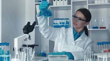 un dedicado hembra científico conduce detallado experimentos con un cautivador azul líquido, meticulosamente analizando datos en un moderno laboratorio lleno con cristalería y avanzado investigación equipo video