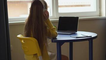 Rear shot of a person in a yellow sweater and headphones using a laptop on a blue table by a window. There is a notepad and a pen near the laptop in a home setting. video