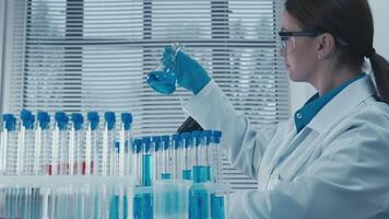 un dedicado científico en laboratorio engranaje examina un vibrante azul solución en un matraz, rodeado por prueba tubos en un lo último investigación laboratorio, demostrando compromiso a científico avance video