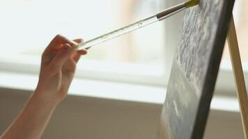 A close-up shot of a hand delicately holding a paintbrush while working on a canvas. The setting appears to be indoors, with natural light subtly illuminating the artistic process. video