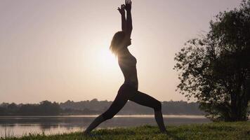 A graceful silhouette of a dedicated yoga practitioner in a serene pose at dawn by shimmering water embodies tranquility and mindfulness in a peaceful natural setting that invites relaxation video