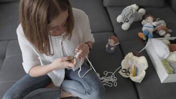 A woman sits on a gray sofa, crocheting with vibrant yarn, surrounded by handmade dolls and soft toys, showcasing her creativity and passion for crafting, embodying relaxation and mindfulness video