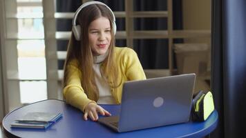 A young girl sits at a round blue table, wearing headphones and using a laptop. She is smiling and appears to be engaged in a call. Notepads and a pencil case are on the table. video