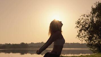 A woman practices yoga at dawn by a tranquil lake, embracing natures stillness. The suns rays illuminate her silhouette, reflecting serenity and inspiring peace and connection to the world video