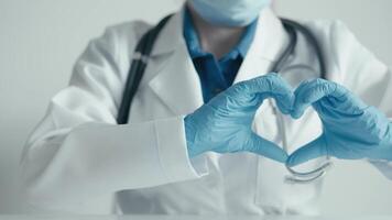 A healthcare professional in a lab coat and gloves forms a heart shape with their hands, symbolizing the compassion and dedication they offer in patient care during challenging times video