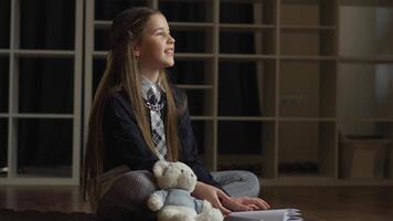 A young schoolgirl sits in a relaxed, cross-legged pose on a wooden floor, dressed in her school attire and accompanied by a teddy bear, seemingly lost in thought as she gazes upwards with a smile. video