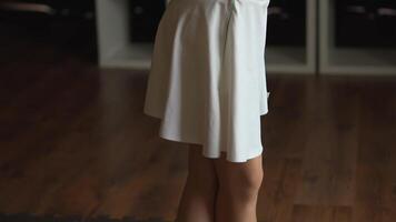 A dancer's legs are depicted in a close-up view, wearing a white dress. The dancer stands poised on a wood and black floor. The lighting creates a minimalist yet elegant atmosphere, emphasizing the grace. video