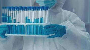 un científico en protector engranaje examina un estante de prueba tubos lleno con vibrante azul soluciones, exhibiendo precisión y avanzado investigación capacidades en un estéril laboratorio ambiente. video