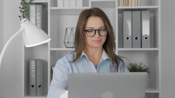 In a vibrant office with greenery and organized files, a focused young woman in glasses smiles while working on her laptop, reflecting a modern, productive, and motivating work environment video