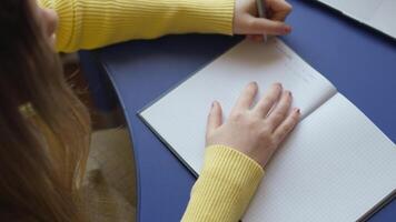 A person is writing in a notebook with graph paper pages. They are wearing a yellow sweater and writing on a blue desk. The focus is on the hand holding the pen and the open notebook. video