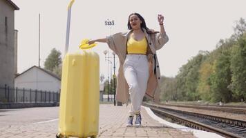 A vibrant young woman in colorful summer attire strolls playfully at a bustling train station with a suitcase, feeling excited and ready for a joyful journey through scenic surroundings video