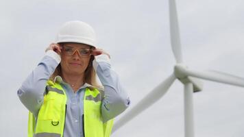 A skilled female engineer adjusts her safety glasses, showcasing professionalism as she stands confidently in front of a massive wind turbine, symbolizing renewable energy and modern innovation video