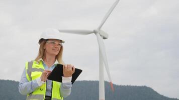 A professional female engineer wearing a helmet and reflective vest reviews data on a tablet while standing in front of a wind turbine, showcasing sustainability technology in action. video